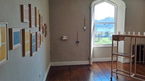 A gallery space with an installation of wooden frames in the centre of the room with framed prints of small text on the wall, a window shows a view of green fields and a lake in the distance.